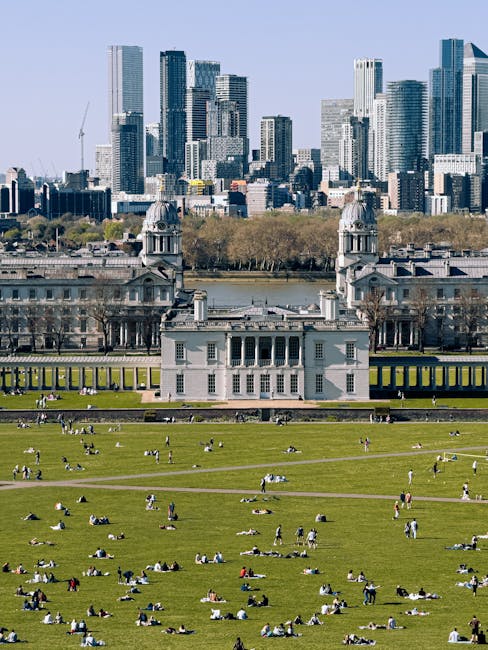 A wide view of a lush green lawn with numerous people relaxing, lounging, and picnicking on the grass in front of the historic Queen's House of Greenwich, which features classical architecture with white walls, columns, and domed towers. Behind the Queen's House, there is a body of water with trees along the shoreline, and a modern city skyline with tall glass skyscrapers under a clear blue sky. The image captures a bright, sunny day with natural light illuminating the landscape, highlighting the contrast between the well-maintained lawns and the urban environment beyond. The scene suggests outdoor leisure and landscape maintenance typical of a historic and outdoor public space managed by professionals like Gardeners Greenwich, serving SE10 homes near Greenwich Park.